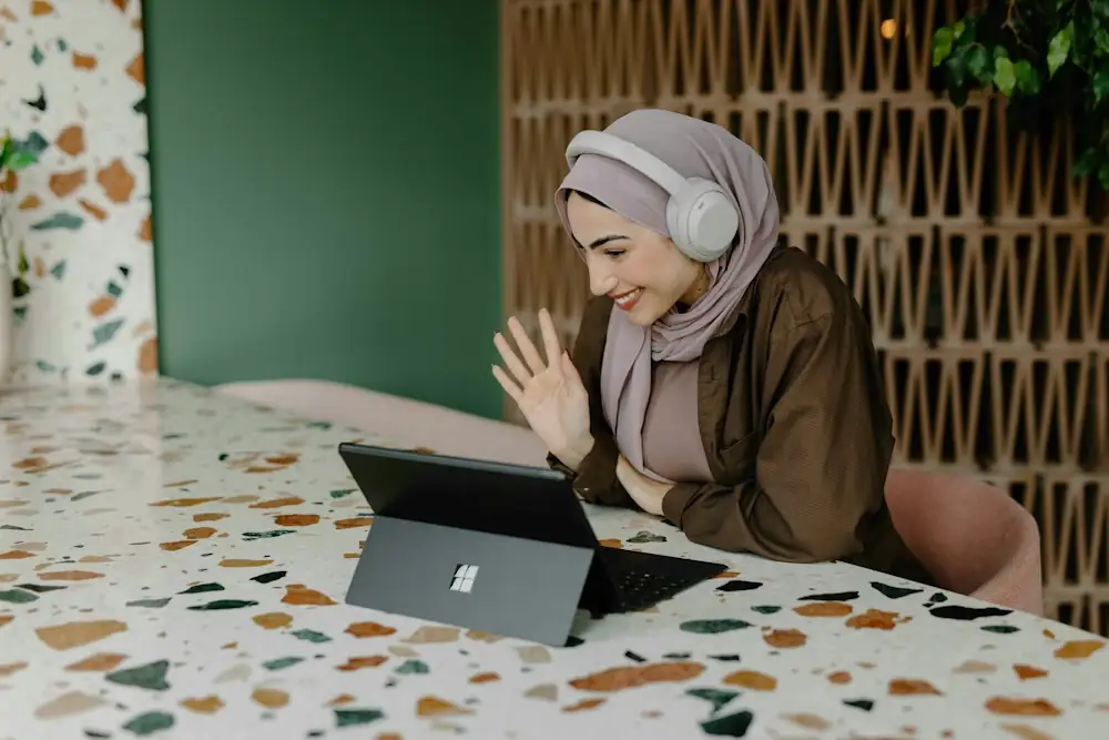 A woman in a hijab smiling and waving during a video call on a tablet